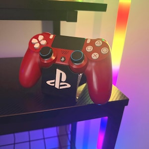 Puede incluir: Un mando rojo de PlayStation 4 con iconos de botones blancos y detalles negros descansa sobre una superficie negra. El mando está encima de una estación de carga negra con el logotipo de PlayStation.