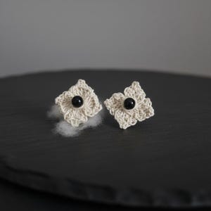 May include: A pair of handmade crochet flower earrings. Each earring features a cream-colored, four-petal flower design with a small, black bead center. The earrings are displayed on a dark surface.