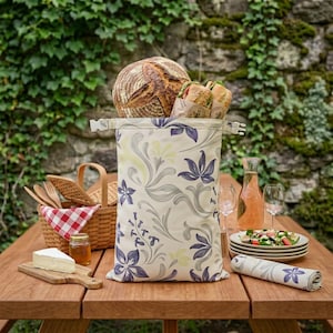 Könnte beinhalten: Eine hohe, rechteckige Stofftasche mit cremefarbenem Hintergrund und einem violett-grauen Blumenmuster, die mit einem Schnallenverschluss versehen ist. Die Tasche ist mit einem runden Brotlaib und zwei in Papier gewickelten Sandwiches gefüllt.