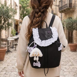 Könnte beinhalten: Ein schwarzer Rucksack mit weißem Gittermuster und weißem Rüschenbesatz an der Klappe. Auf der Klappe befindet sich eine Stickerei mit dem Text I heart und einem Flügelsymbol. Ein weißer Plüsch-Pilzanhänger hängt seitlich an einem weißen Etikett.