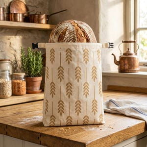 Könnte beinhalten: Ein beiger Brotbeutel mit einem Weizenährenmuster enthält ein Brot. Die Tasche hat einen schwarzen Schnallenverschluss und steht auf einer Holztheke. Eine Kupferkanne und andere Küchenartikel befinden sich im Hintergrund.