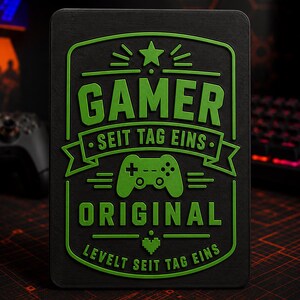May include: A rectangular black plaque with raised neon green text and graphics. The design features a star, a game controller icon, and a pixelated heart. The text reads: GAMER SEIT TAG EINS ORIGINAL LEVELT SEIT TAG EINS in a bold, layered style.