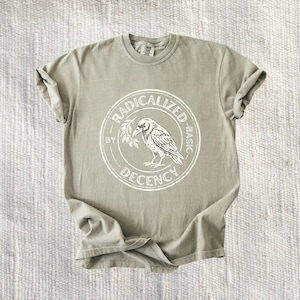 Może przedstawiać: Jasnooliwkowy t-shirt z krótkim rękawem z okrągłą białą grafiką na klatce piersiowej. Wzór przedstawia kruka siedzącego na gałązce oliwnej, otoczonego obwódką z napisem "RADICALIZED BY DECENCY" wykonanym pogrubioną czcionką.