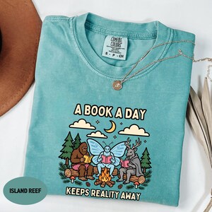 May include: A teal short-sleeve t-shirt featuring a graphic of a Bigfoot, a fairy, and a deer sitting around a campfire reading books. The text reads A BOOK A DAY KEEPS REALITY AWAY. The shirt has a Comfort Colors label and a crew neckline.