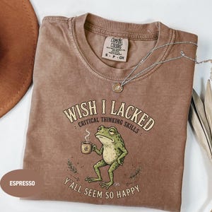 May include: An espresso-coloured t-shirt featuring a graphic of a green frog holding a steaming mug. The text reads: WISH I LACKED CRITICAL THINKING SKILLS Y'ALL SEEM SO HAPPY. The shirt has a crew neckline and a Comfort Colors brand label inside.