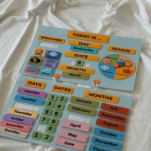 K&ouml;nnte beinhalten: Ein p&auml;dagogischer Filz-Kalender mit Abschnitten f&uuml;r Wetter, Tag, Datum, Monat und Jahreszeit. Enth&auml;lt Etiketten f&uuml;r Wochentage, Zahlen und Monate. Der Text "TODAY IS..." ist oben sichtbar. Der Kalender befindet sich auf einer wei&szlig;en Oberfl&auml;che.