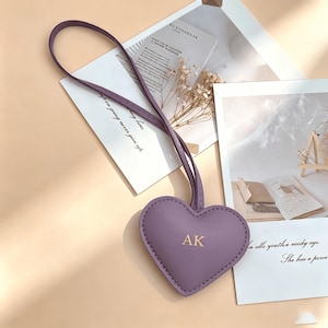 May include: A lavender heart-shaped keychain with the initials "AK" in gold. The keychain has a matching strap and is set against a backdrop of stationery and dried flowers. The text "tap on going never give up" is visible.