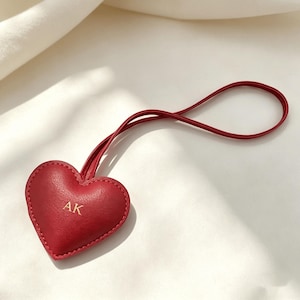 Puede incluir: Un llavero de cuero rojo en forma de corazón con las iniciales "AK" en letras doradas. El llavero tiene un lazo para colgar. El corazón está cosido alrededor de los bordes y se encuentra sobre una superficie blanca.