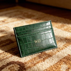 May include: A dark green rectangular card holder featuring a textured crocodile skin pattern and gold embossed initials WD on the front. The slim accessory includes multiple horizontal slots for organizing cards and is finished with tonal stitching.