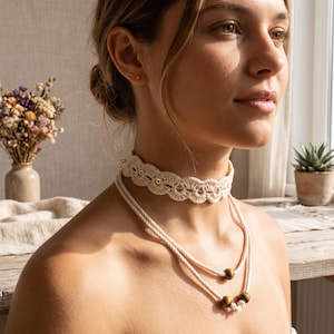Op de afbeelding: Een gelaagde kettingenset bestaande uit een crèmekleurige gehaakte kanten choker met kleine goudkleurige kralen in het schulppatroon, gecombineerd met een dubbele koordketting versierd met drie gladde, donkerbruine houten kralen.