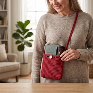 May include: A deep red crochet crossbody bag featuring a single wooden button closure at the top. The bag has a long, matching red crocheted strap and contains a dark green notebook, a pen, and a makeup brush inside the main compartment.