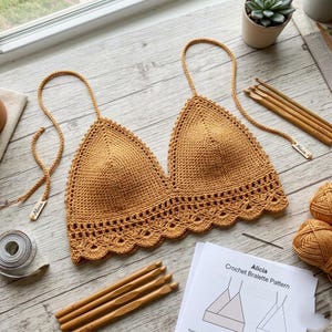 May include: A handmade, mustard-colored crochet bralette with a scalloped edge and adjustable straps. The bralette is displayed with crochet hooks, yarn, and a pattern sheet. The pattern sheet is labeled "Alicia Crochet Bralette Pattern".