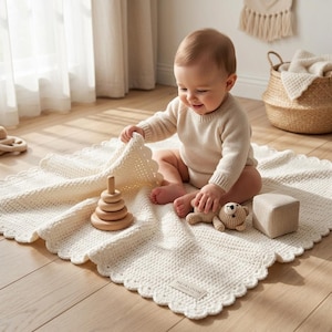 Pode incluir: Uma manta de bebé em croché cor de creme com rebordo recortado e uma pequena etiqueta retangular com a inscrição HEIRLOOM. A manta apresenta um padrão de ponto texturado e é exibida com um brinquedo de empilhar, um urso e um cubo.