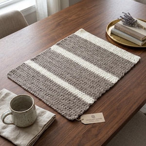 May include: A rectangular crochet placemat featuring alternating horizontal stripes of taupe and cream yarn. A small beige paper tag attached to the corner displays the text CROCHET HOME ARTISANAL QUALITY in black ink with a small yarn ball icon.