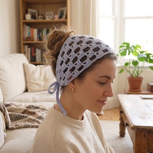 May include: A light lavender crochet headscarf featuring an openwork grid pattern. The headscarf is triangular in shape and includes thin crocheted ties at the back to secure it in place, providing a lightweight and textured accessory for the hair.