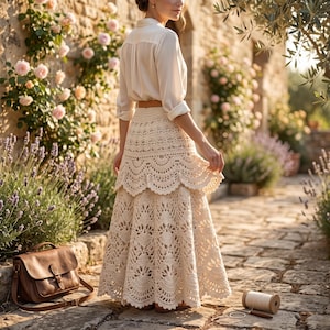 May include: A long, cream-coloured crochet skirt featuring an intricate, tiered scalloped pattern and a delicate openwork design. The skirt is paired with a plain, long-sleeved, light-coloured button-down shirt and a simple brown leather belt.