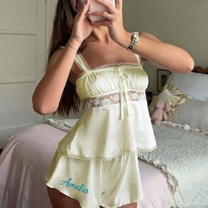 May include: A pale yellow satin pajama set consisting of a camisole and shorts. The camisole features a square neckline, thin straps, and a horizontal band of white floral lace with a tie detail. The shorts are embroidered with the name Amelia.