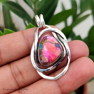 May include: A pendant featuring a triangular pink stone with gold and blue veining, encased in a polished silver frame with overlapping, twisted metal bands. The piece includes a silver bail at the top for attachment to a chain or cord.