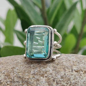 May include: A silver ring featuring a large, rectangular, faceted light blue gemstone set in a bezel. The band consists of three thin, parallel silver wires that connect to the sides of the setting, which is accented with small silver spheres.