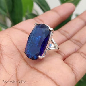 Könnte beinhalten: Ein Silberring mit einem großen, länglichen, oval facettierten tiefblauen Edelstein, der von vier Krappen gehalten wird. Die Ringschiene ist in zwei dünne Silbersegmente unterteilt, die mit der Fassung verbunden sind. Der Text Kingman Jewelry Shop ist sichtbar.