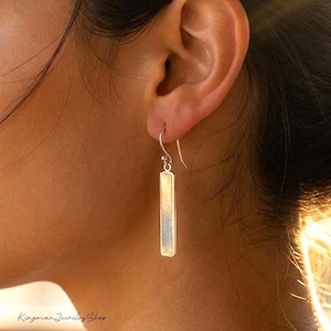 May include: A silver-toned drop earring featuring a slender, rectangular bar pendant suspended from a curved hook wire. The metal surface has a subtle, brushed texture that catches the light, creating a clean and simple vertical design.