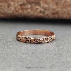May include: A copper-toned metal ring featuring an embossed floral and vine pattern around the entire band. The surface of the ring has a textured, oxidized finish that highlights the intricate botanical details carved into the metal band.