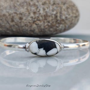 May include: A silver cuff bracelet featuring a central oval-shaped stone with a distinct black and white marbled pattern. The metal band is smooth and polished, with a gap at the back for adjustment. The text KingmanJewelryShop is visible at the bottom.