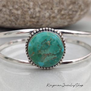 Peut inclure: Un bracelet manchette en argent avec une pierre turquoise ronde et polie présentant des veines de matrice brune. La pierre est sertie dans une bordure en argent perlée. Le bracelet se compose de deux fils d'argent parallèles. Texte : Kingman Jewelry Shop.
