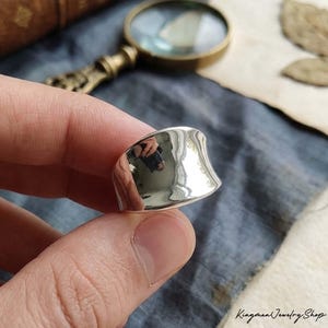 Könnte beinhalten: Ein polierter Silberring mit geschwungenem, asymmetrischem Design. Der Ring reflektiert die Umgebung und zeigt seine glatte, reflektierende Oberfläche. Der Ring wird von den Fingern einer Person gehalten. Der Shop-Name "Kingman Jewelry Shop" ist sichtbar.