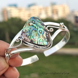 May include: A silver cuff bracelet featuring a large, triangular abalone shell inlay with iridescent swirls of green, blue, and purple. The metal band has a polished finish with decorative wire scrollwork accents on either side of the central stone.