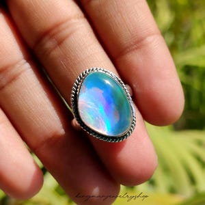 May include: A silver ring with a large, oval-shaped opal gemstone. The opal displays a vibrant play of color, with hues of blue, green, and purple. The ring has a twisted rope-like border. The text "kingmanjewelryshop" is at the bottom.