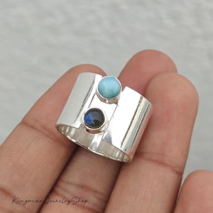 May include: A wide silver band ring featuring two vertically aligned round cabochon gemstones. The top stone is a smooth, opaque light blue, while the bottom stone is a dark, iridescent gray. The text KingmanJewelryShop is visible.