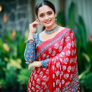 May include: A red saree featuring a repeating blue and white floral block print pattern, paired with a matching long-sleeved blouse. The ensemble includes a silver-toned choker necklace and a silver-toned bracelet on the wrist.