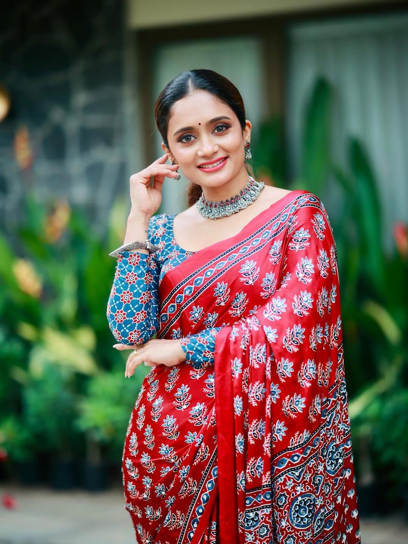 May include: A red saree featuring a repeating blue and white floral block print pattern, paired with a matching long-sleeved blouse. The ensemble includes a silver-toned choker necklace and a silver-toned bracelet on the wrist.