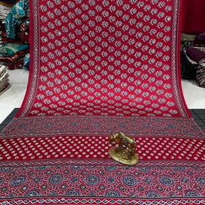 May include: A vibrant red fabric featuring a repeating white floral pattern and small reflective accents. The lower section displays a complex border with dark blue and white geometric motifs, complemented by a small gold-toned cow figurine.