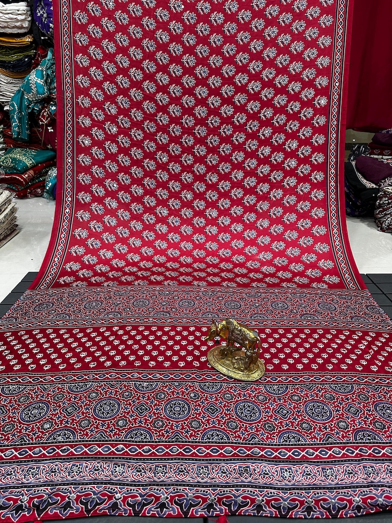 May include: A vibrant red fabric featuring a repeating white floral pattern and small reflective accents. The lower section displays a complex border with dark blue and white geometric motifs, complemented by a small gold-toned cow figurine.