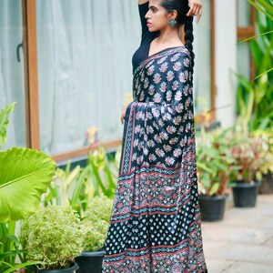 May include: A black saree featuring a repeating pattern of small floral motifs and geometric borders in shades of red, white, and blue. The garment is paired with a solid black short-sleeved blouse, creating a cohesive look with intricate detailing.