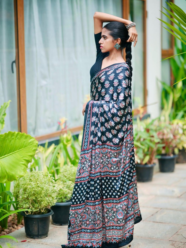 May include: A black saree featuring a repeating pattern of small floral motifs and geometric borders in shades of red, white, and blue. The garment is paired with a solid black short-sleeved blouse, creating a cohesive look with intricate detailing.