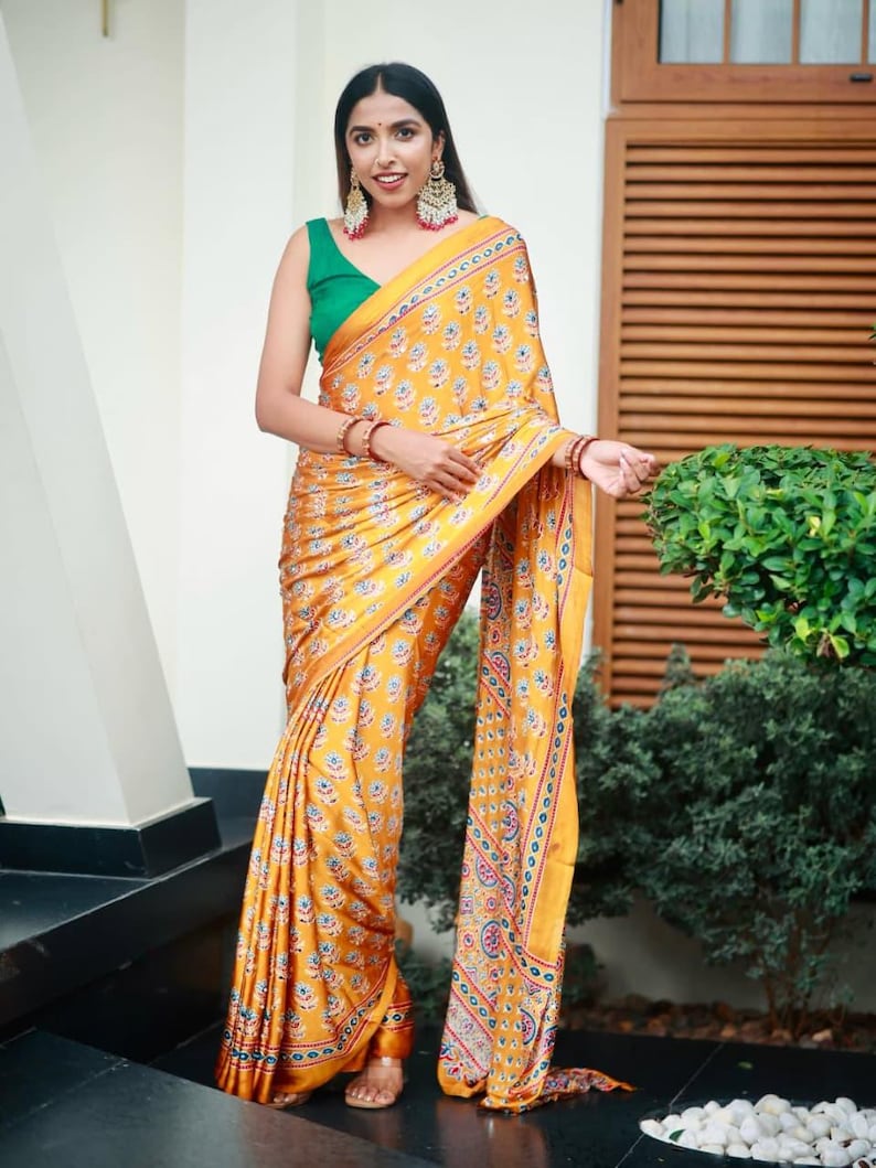 May include: A mustard yellow saree featuring a repeating floral pattern in blue, red, and white, paired with a sleeveless emerald green blouse. The saree includes a wide decorative border with intricate geometric and floral motifs in matching colors.