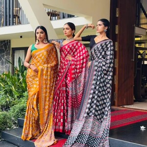 May include: Three sarees featuring intricate floral block prints in mustard yellow, vibrant red, and deep black. Each garment is draped in a traditional style, showcasing detailed patterns, contrasting borders, and flowing fabric that reaches the ground.