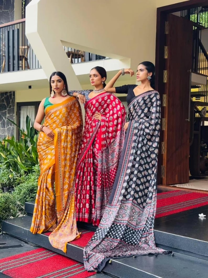 May include: Three sarees featuring intricate floral block prints in mustard yellow, vibrant red, and deep black. Each garment is draped in a traditional style, showcasing detailed patterns, contrasting borders, and flowing fabric that reaches the ground.