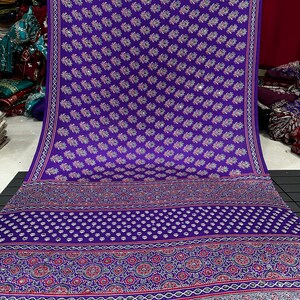 May include: A vibrant purple fabric featuring a repeating pattern of small floral motifs interspersed with reflective circular accents. The lower section displays a wide, intricate border with geometric shapes, dots, and floral designs in red and white.