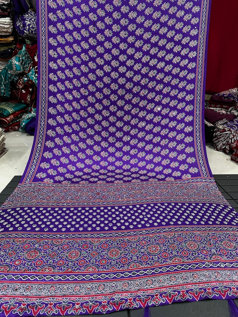 May include: A vibrant purple fabric featuring a repeating pattern of small floral motifs interspersed with reflective circular accents. The lower section displays a wide, intricate border with geometric shapes, dots, and floral designs in red and white.