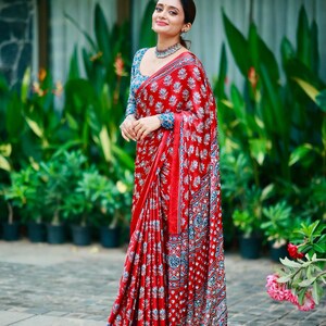May include: A vibrant red saree featuring a repeating white and blue floral block print pattern. The garment includes a matching long-sleeved blouse with a similar intricate blue and white botanical design, paired with a silver-toned choker necklace.