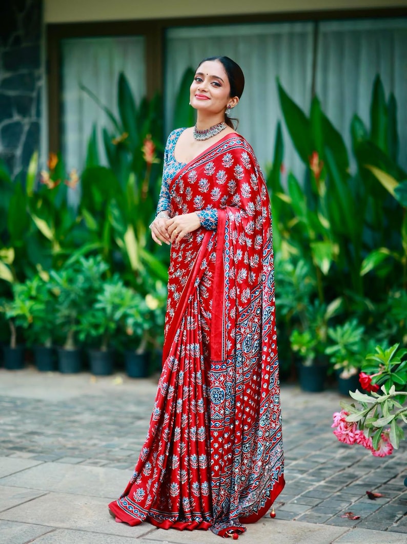 May include: A vibrant red saree featuring a repeating white and blue floral block print pattern. The garment includes a matching long-sleeved blouse with a similar intricate blue and white botanical design, paired with a silver-toned choker necklace.
