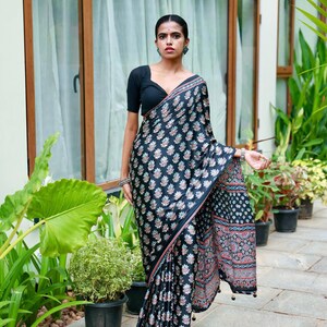 May include: A black saree featuring a repeating floral block print pattern in shades of blue, white, and red. The garment includes a solid black short-sleeved blouse with a V-neckline and a decorative border along the edge of the fabric.