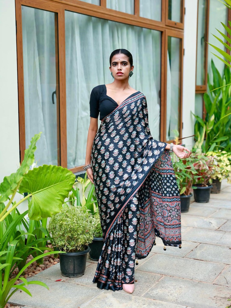 May include: A black saree featuring a repeating floral block print pattern in shades of blue, white, and red. The garment includes a solid black short-sleeved blouse with a V-neckline and a decorative border along the edge of the fabric.