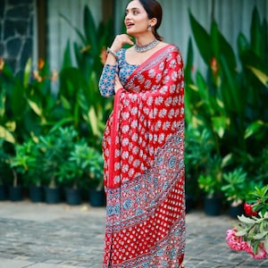 May include: A red saree featuring a repeating white floral block print pattern with a blue and white geometric border. The matching long-sleeved blouse displays a blue base with a white floral pattern, complemented by a silver-toned choker necklace.