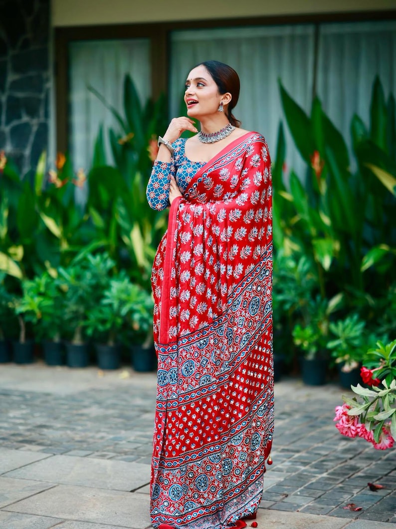 May include: A red saree featuring a repeating white floral block print pattern with a blue and white geometric border. The matching long-sleeved blouse displays a blue base with a white floral pattern, complemented by a silver-toned choker necklace.