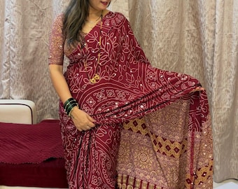 May include: A deep maroon saree featuring intricate white bandhani-style patterns and small reflective mirror embellishments. The garment includes a matching blouse with short sleeves and a wide, decorative border with geometric gold-toned motifs.